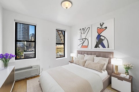 A bedroom with a large bed, a painting on the wall, and a view of a cityscape through the window.